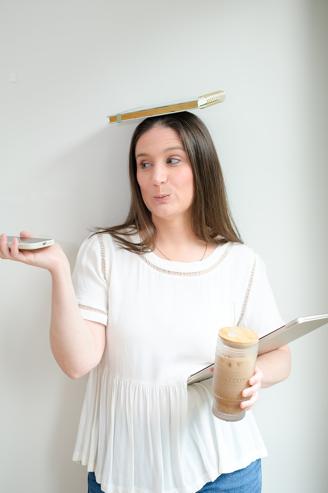 Chelsea balancing a notebook on her head while holding a phone, laptop and iced coffee