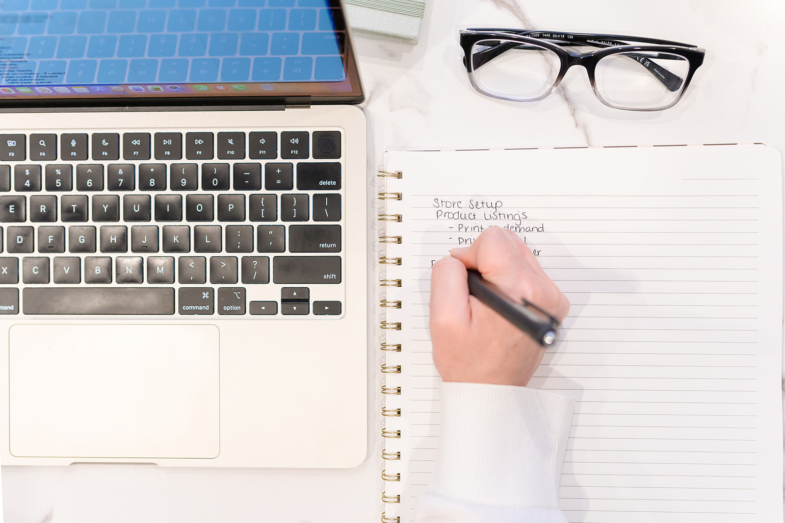 Hand writing a store setup checklist in a notebook beside a laptop